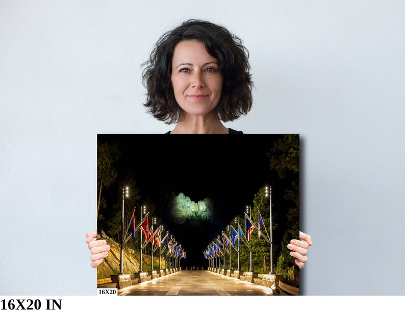 May include: A black and white photograph of Mount Rushmore at night with a row of flags lining the road leading up to the monument. The flags are lit up with string lights.