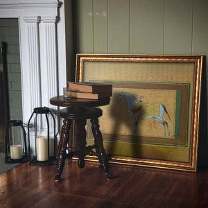 May include: A dark wood stool with a round top and three legs sits in front of a gold framed picture of a white horse with a rider. The picture is hanging on a green wall. Two lanterns are on the floor to the left of the stool.