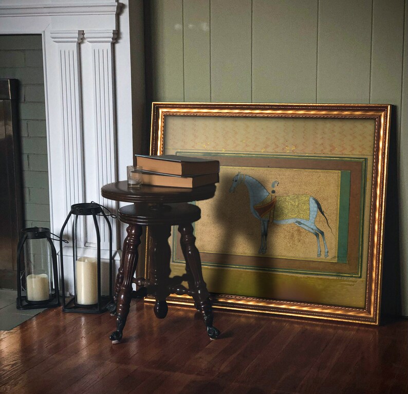 May include: A dark wood stool with a round top and three legs sits in front of a gold framed picture of a white horse with a rider. The picture is hanging on a green wall. Two lanterns are on the floor to the left of the stool.