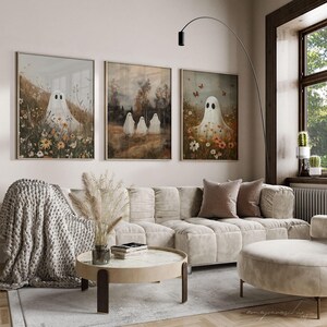 May include: Three framed art prints featuring white ghosts in a field of flowers. The prints are hung on a white wall above a white sofa with a brown throw blanket and a coffee table.