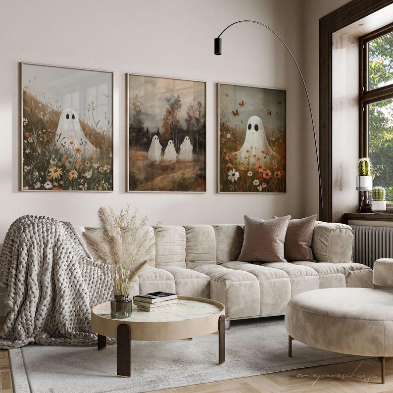 May include: Three framed art prints featuring white ghosts in a field of flowers. The prints are hung on a white wall above a white sofa with a brown throw blanket and a coffee table.