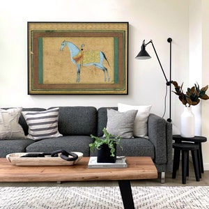 May include: A gray sofa with pillows and a wooden coffee table in front of it. A framed painting of a blue horse with a rider is hanging on the wall above the sofa. A black lamp with a black shade is mounted on the wall to the right of the painting.