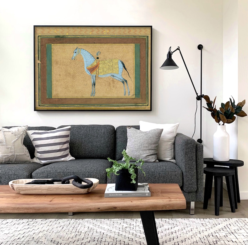 May include: A gray sofa with pillows and a wooden coffee table in front of it. A framed painting of a blue horse with a rider is hanging on the wall above the sofa. A black lamp with a black shade is mounted on the wall to the right of the painting.