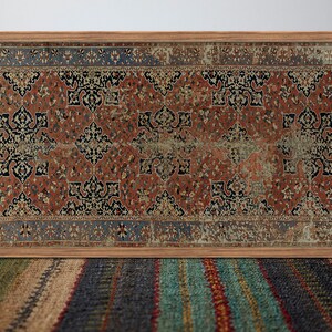 K&ouml;nnte beinhalten: Ein gerahmter Vintage-Teppich mit einem geometrischen Muster in Rot, Blau und Schwarz. Der Teppich befindet sich in einem Holzrahmen und h&auml;ngt an einer wei&szlig;en Wand.