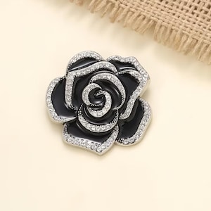 May include: A black and silver rose-shaped brooch. The brooch features a black enamel base with a silver outline and is encrusted with clear crystals. The rose design is detailed and elegant, suitable for formal wear.
