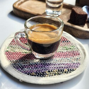 May include: A small glass cup of espresso coffee on a colorful woven coaster. The coffee is dark brown and has a light brown crema on top.