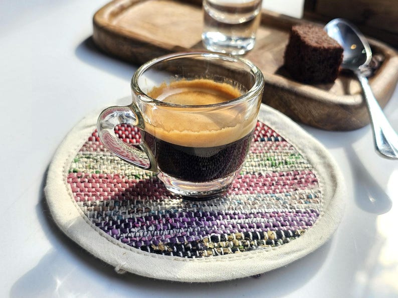 May include: A small glass cup of espresso coffee on a colorful woven coaster. The coffee is dark brown and has a light brown crema on top.