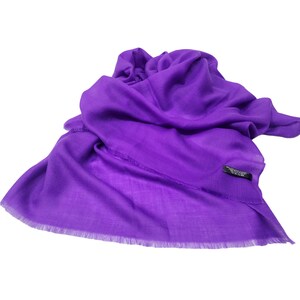Puede incluir: Una bufanda morada vibrante con un borde deshilachado. La bufanda está drapeada, mostrando su textura suave y color. Una pequeña etiqueta negra es visible. Este accesorio es adecuado para diversas ocasiones.