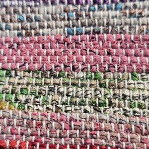 May include: Close-up of a woven rug with a multi-colored striped pattern. The rug is made of different colored fabrics, including pink, green, white, and blue.