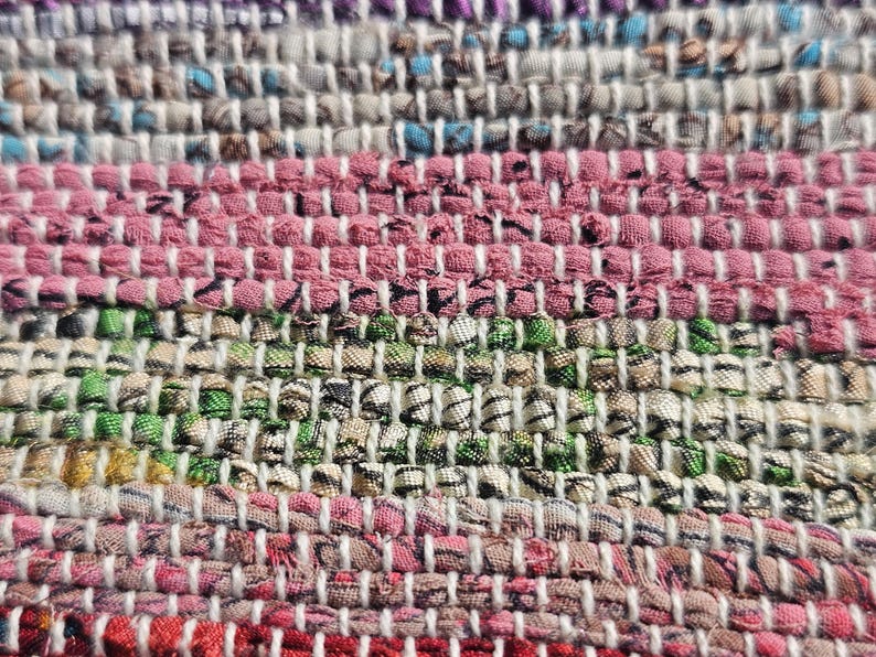 May include: Close-up of a woven rug with a multi-colored striped pattern. The rug is made of different colored fabrics, including pink, green, white, and blue.