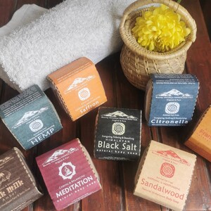 May include: Assortment of natural Himalayan body soap bars with various scents, including Hemp, Saffron, Citronella, Frankincense, Black Salt, Meditation, Sandalwood, and Yak Milk Amber. The soaps are packaged in brown paper with black and white text and a mountain graphic.
