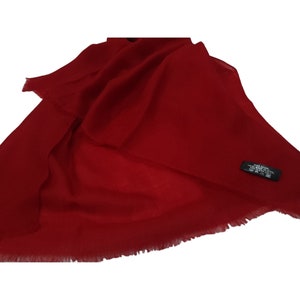 Puede incluir: Una bufanda de cachemira roja con borde de flecos. La bufanda tiene una etiqueta negra con el texto "100% CASHMERE HAND-WOVEN IN ITALY DRY CLEAN ONLY".