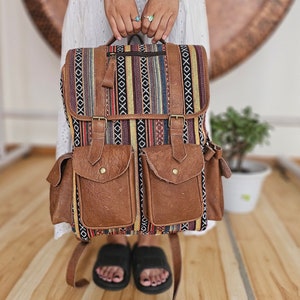 Pode incluir: Uma mochila de couro marrom com uma aba de tecido tecido colorido. A mochila tem dois bolsos grandes na frente e alças ajustáveis.