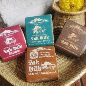 May include: Four bars of handmade soap with the words "Yak Milk" on the packaging. The soaps are in a woven basket with a white towel and a small basket of yellow flowers in the background. The soap scents are Rose, Fruity, Amber, and Sandalwood.
