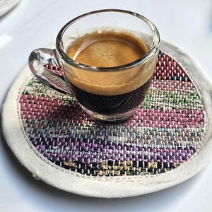 May include: A small glass cup of espresso coffee on a colorful woven coaster. The coaster is made of different colored fabrics and has a white border.