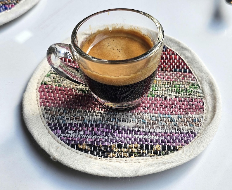 May include: A small glass cup of espresso coffee on a colorful woven coaster. The coaster is made of different colored fabrics and has a white border.
