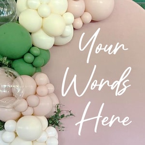 May include: A pink circular backdrop with the text "Your Words Here" in white script. The backdrop is decorated with clusters of balloons in white, light pink, and green. Greenery is also visible.