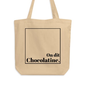 May include: A beige tote bag with black handles and a black square on the front. The square contains the text "On dit Chocolatinee."