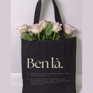 May include: Black tote bag with white text that reads "Ben l&agrave;." The bag has a quote in French about the meaning of the phrase. The bag is filled with pink roses.