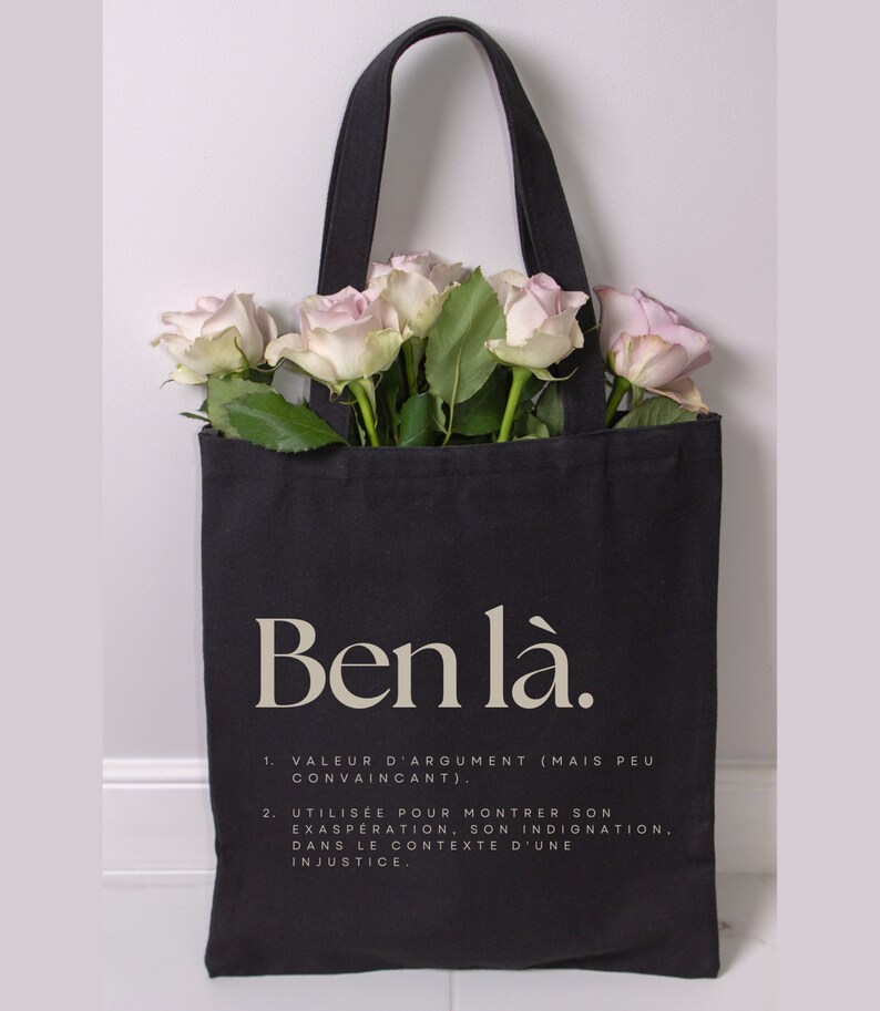 May include: Black tote bag with white text that reads "Ben l&agrave;." The bag has a quote in French about the meaning of the phrase. The bag is filled with pink roses.