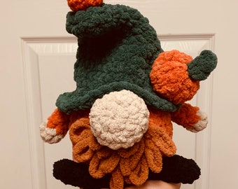 May include: A handmade, plush gnome with a green hat and orange pumpkin accents. The gnome has a white beard, orange body, and black shoes. The gnome is a fall-themed decoration.