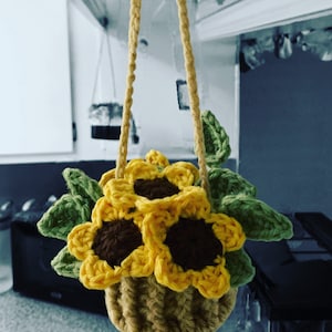 Könnte beinhalten: Ein gehäkelter Hängekorb mit drei gelben Sonnenblumen und grünen Blättern. Der Korb ist aus braunem Garn gefertigt und hat eine gewebte Textur.