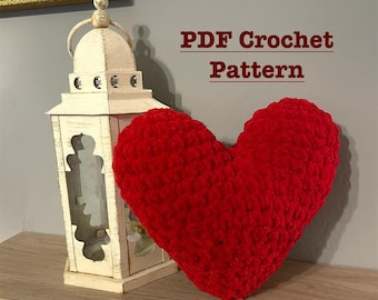 May include: A red crocheted heart-shaped pillow. The pillow is on a wooden surface next to a white lantern. The text "PDF Crochet Pattern" is written in red letters on a gray background.