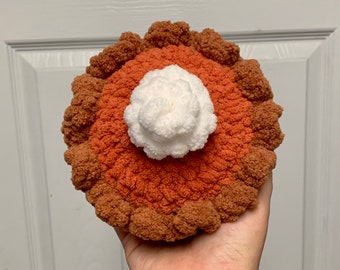 May include: A crocheted pumpkin pie with a white whipped cream topping. The pie is brown and orange and has a textured surface.