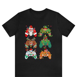 May include: Black t-shirt featuring six Christmas-themed video game controller designs. The controllers are decorated as Santa Claus, an elf, a reindeer, a Christmas tree, a snowman, and a gingerbread man, all adorned with colorful string lights.