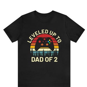Può includere: Maglietta nera con una grafica retrò di un controller per videogiochi e il testo "LEVELED UP TO DAD OF 2".