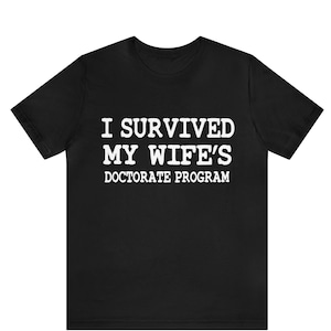May include: Black t-shirt with the white text: "I SURVIVED MY WIFE'S DOCTORATE PROGRAM". The crew neck shirt is made of a soft material. The text is centered on the front of the shirt, making a bold statement.