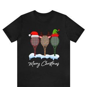 Może przedstawiać: Czarny T-shirt z świątecznym wzorem przedstawiającym trzy rakiety tenisowe ozdobione kratą, nadrukiem lamparta i zielono-czerwonymi wzorami. Rakietki są ozdobione czapkami Świętego Mikołaja, rogami renifera i czapkami elfów. Tekst "Merry Christmas" jest nadrukowany pod rakietkami.