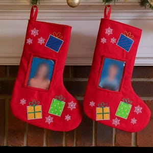 May include: Two red Christmas stockings with photo inserts, decorated with embroidered snowflakes and gift boxes. Each stocking has a loop for hanging. Festive holiday decor.