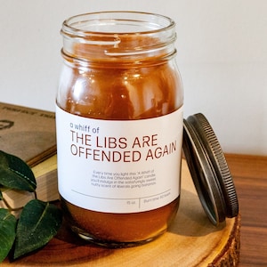 May include: A brown glass jar candle with a white label that reads "A whiff of THE LIBS ARE OFFENDED AGAIN". The label also includes a description of the scent. The candle is sitting on a wooden surface with a book and green leaves.