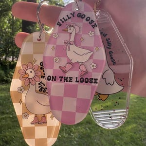 May include: Three novelty keychains with goose designs. One keychain is yellow with a daisy and the words "Good Vibe". The second keychain is pink checkered with a goose wearing a hat and boots, and the words "Silly Goose On The Loose". The third keychain is clear with a goose.