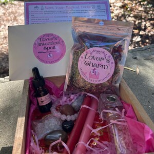 May include: A wooden box filled with various items for a love spell. Includes pink candles, a bottle of oil, a bag of herbs, crystals, and a bracelet. The box also contains a card that reads "Meet Your Magical Tools".