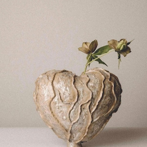 Pode incluir: Um vaso em forma de coração, texturizado, em tons de bege e cinza, com duas flores secas com folhas verdes. O vaso tem um design único e está sobre uma superfície branca.