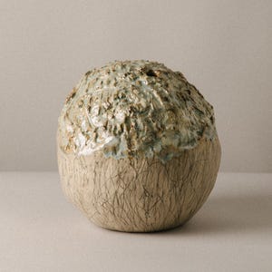 Könnte beinhalten: Eine kugelförmige Keramikvase mit einem strukturierten, unebenen oberen Bereich, der eine glänzende, hellblaue und braune Glasur aufweist. Der untere Teil der Vase hat eine glatte, matte beige Oberfläche mit feinen, dunklen, sich kreuzenden Linien.