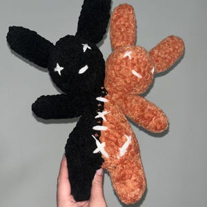 May include: A handmade plush bunny toy, half black and half orange, with white cross-stitch details. The bunny has long ears and is made of a soft, textured material. The two halves are sewn together with white thread.
