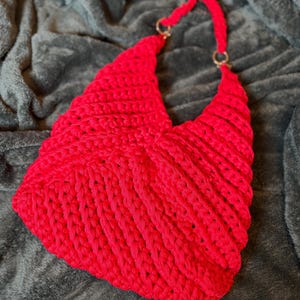 May include: A vibrant red crocheted handbag with a unique, textured design. The bag features a structured shape with a handle attached by gold-colored rings. The handmade bag is made of a thick, soft yarn.