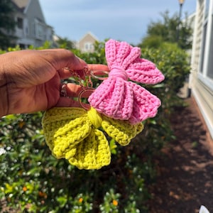 May include: Two crochet bow keychains attached to a gold-toned metal ring. One bow is a vibrant lime green color, and the other bow is a bright bubblegum pink color. Both bows feature a textured, ribbed yarn construction with a central knot detail.