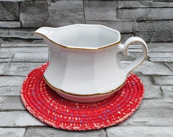 May include: A white ceramic gravy boat with a gold trim sits on a red and white woven coaster.