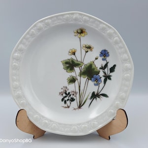 May include: White ceramic plate with a floral design featuring yellow and blue flowers, green leaves, and a decorative rim. The plate is displayed on a wooden stand. The plate's diameter is approximately 8 inches.
