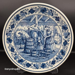 May include: A decorative ceramic plate with a blue and white pastoral scene. The plate features a detailed illustration of people working with cows and a barrel, surrounded by a floral border. The plate is on a clear stand.