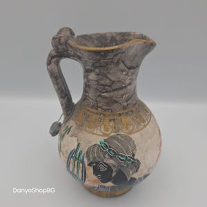 May include: A decorative ceramic jug with a handle and spout. The jug features a textured grey top and a cream-coloured body with gold accents and a black and green illustration of a person.