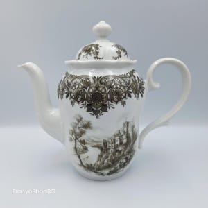 Puede incluir: Tetera de porcelana blanca con un patrón floral decorativo alrededor de la parte superior y en la tapa. La tetera presenta un diseño de paisaje con un castillo y árboles. El asa y el pico son curvos.