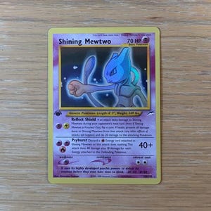 May include: A Shining Mewtwo Pokémon trading card with a purple and gold border. The card features a blue Mewtwo illustration with the text "Shining Mewtwo" and "70 HP". The card also includes attack details and other game information.
