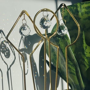 May include: Three gold metal decorative stands with crystal prisms hanging from them. The stands are in a gray pot with white stones.