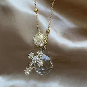 May include: A gold chain necklace with a crystal pendant, a pearl-embellished cross charm, and a white flower charm.