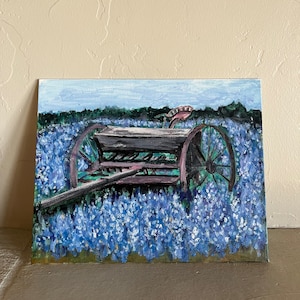 May include: A painting of a vintage farm wagon in a field of bluebonnet flowers. The wagon is brown and has two large wheels. The flowers are a vibrant blue and are in full bloom.